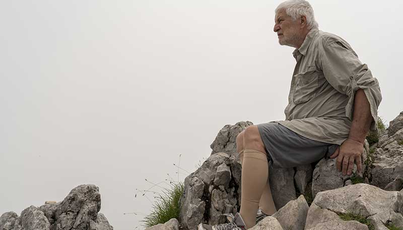 older grey-haired man wearing compression socks sitting on a rock outcropping
