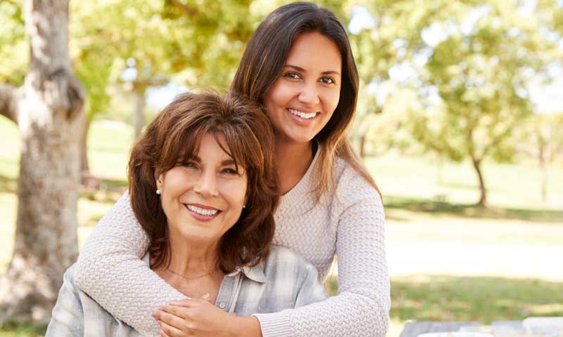 Outside in a park, a standing daughter has her arms gently around her sitting mother