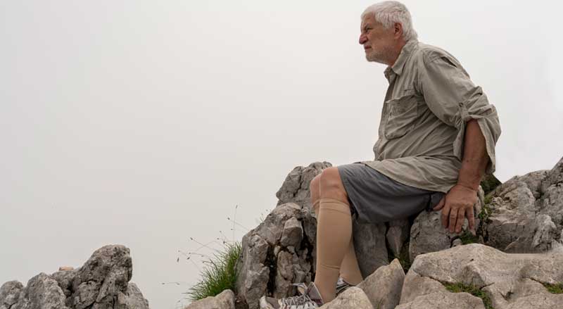 An older man wearing shorts and compression socks sitting on rocks outdoors